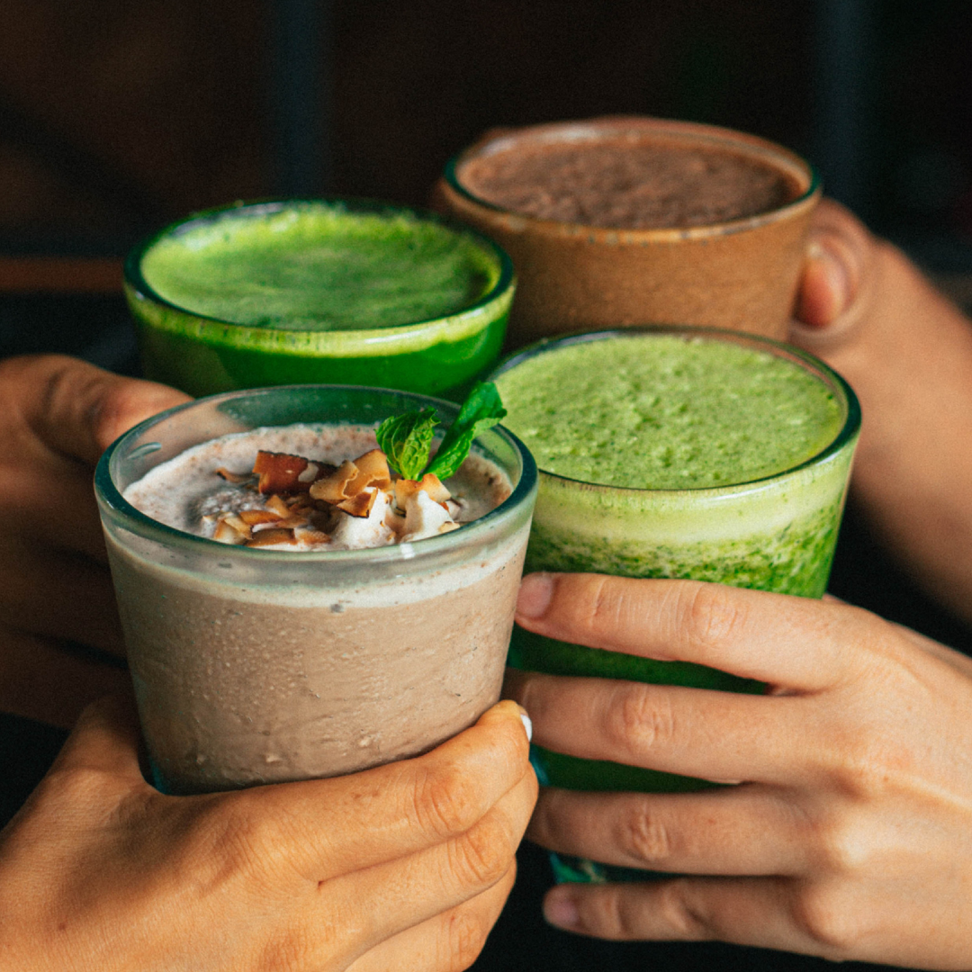 Vier Hände halten verschiedene Café NINI Getränke zusammen, grüne Matcha Drinks und cremige Smoothies in Gläsern, lebhaftes Lifestyle Bild mit frischen Zutaten