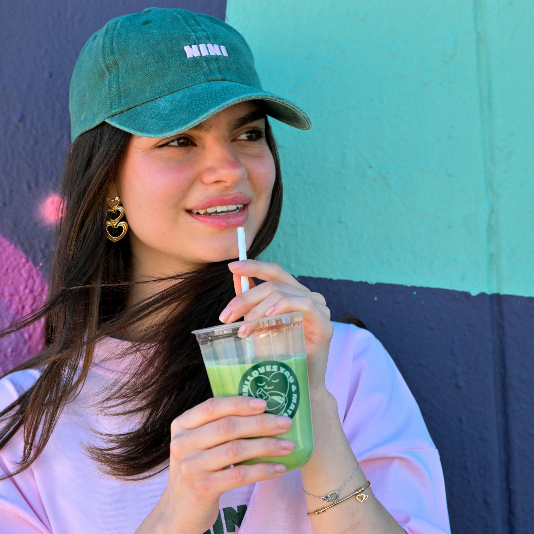NINI Matcha Cap in Grün, getragen von Frau mit Iced Matcha Latte vor bunter Wand