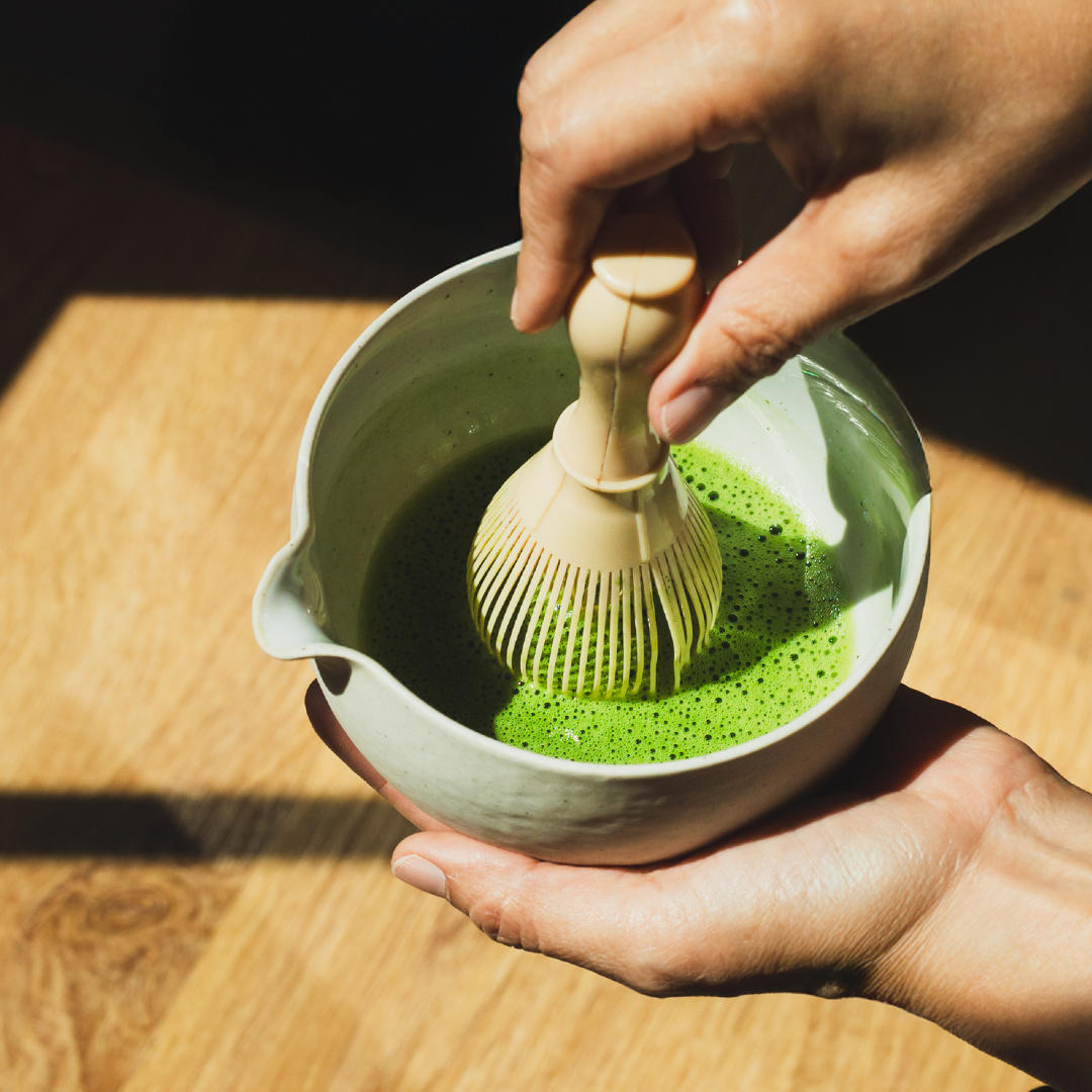 Matcha wird mit einem Bambus Chasen in einer Keramik Matchaschale aufgeschäumt, lebendiges Grün des NINI Matcha Ceremonial Grade sichtbar, authentische Zubereitung auf Holzunterlage