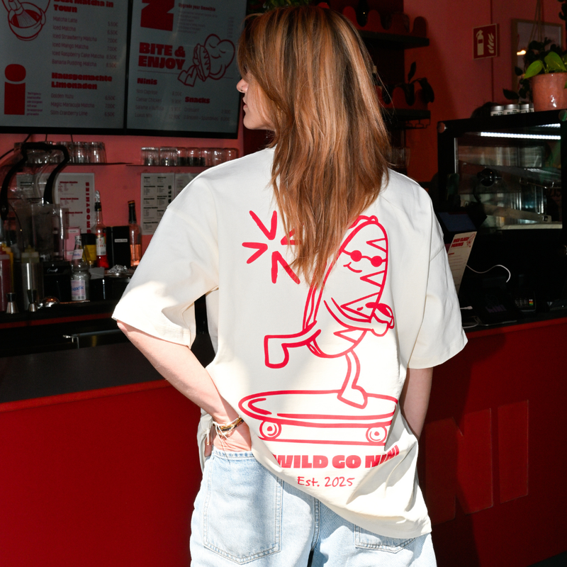 Frau trägt Café NINI Oversized T-Shirt in Creme mit rotem „Go Wild Go NINI Est. 2025" Skater Backprint, Nahaufnahme des Rückenprints im Café NINI