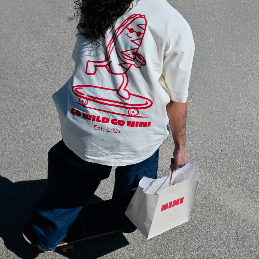 Person trägt Café NINI Oversized T-Shirt in Creme mit rotem „Go Wild Go NINI Est. 2025" Skater Backprint und hält eine weiße NINI Papiertragetasche, Vogelperspektive auf Asphalt mit Skateboard