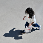Frau fährt Skateboard und trägt Café NINI Oversized T-Shirt in Creme mit rotem NINI Frontprint, hält weiße NINI Tragetasche, Vogelperspektive mit Schatten auf Asphalt