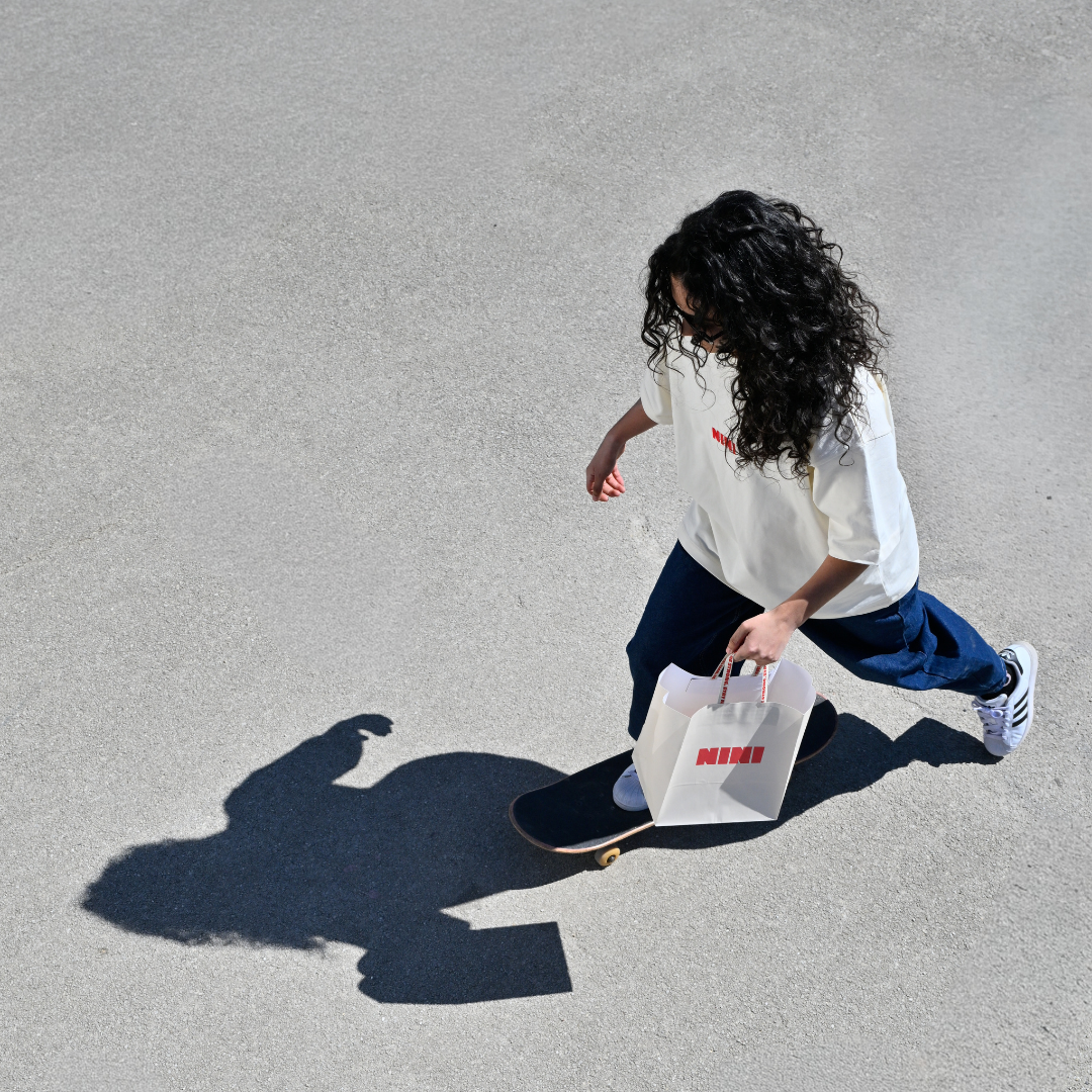 Frau fährt Skateboard und trägt Café NINI Oversized T-Shirt in Creme mit rotem NINI Frontprint, hält weiße NINI Tragetasche, Vogelperspektive mit Schatten auf Asphalt