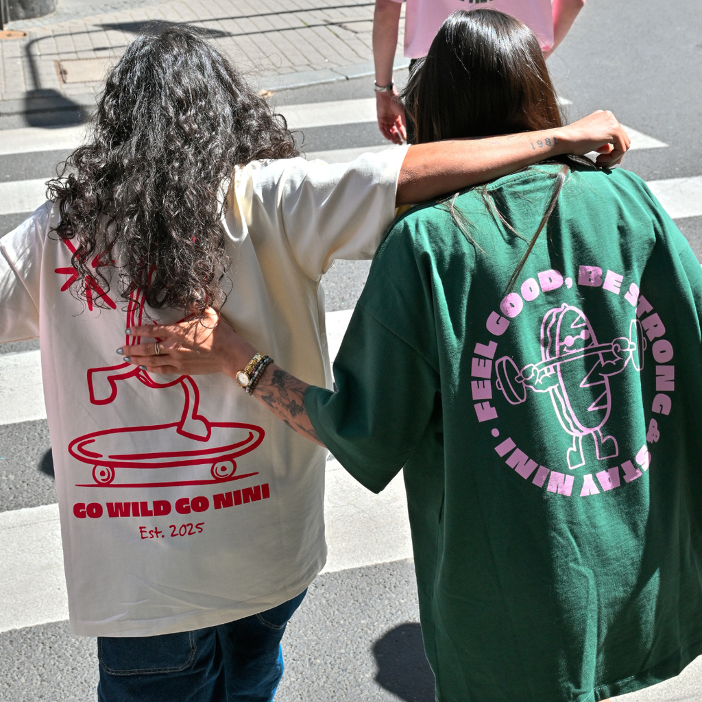 Zwei Personen laufen Arm in Arm und tragen Café NINI Oversized T-Shirts, creme mit rotem „Go Wild Go NINI Est. 2025" Backprint und grün mit pinkem „Feel Good Be Strong Stay NINI" Runddruck, urbaner Streetstyle