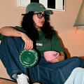 Frau sitzt entspannt und trägt Café NINI Oversized T-Shirt in Grün mit NINI Frontprint und grüne NINI Cap, hält pinke Café NINI Keramiktasse und grüne Untertasse mit „Feel Good Be Strong Stay NINI" Schrift, gemütliches Indoor Lifestyle Lookbook
