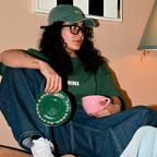 Frau sitzt entspannt und trägt Café NINI Oversized T-Shirt in Grün mit NINI Frontprint und grüne NINI Cap, hält pinke Café NINI Keramiktasse und grüne Untertasse mit „Feel Good Be Strong Stay NINI" Schrift, gemütliches Indoor Lifestyle Lookbook