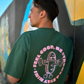 Mann trägt Café NINI Oversized T-Shirt in Grün mit pinkem „Feel Good Be Strong Stay NINI" Runddruck auf dem Rücken, Nahaufnahme des Backprints vor Graffiti-Wand