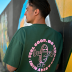 Mann trägt Café NINI Oversized T-Shirt in Grün mit pinkem „Feel Good Be Strong Stay NINI" Runddruck auf dem Rücken, Nahaufnahme des Backprints vor Graffiti-Wand