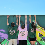 Drei Personen stehen vor einer Graffiti-Wand und tragen Café NINI Oversized T-Shirts, zwei grüne Shirts mit „Be Good Be Strong Stay NINI" Backprint und ein pinkes Shirt mit „Matcha Loves You & NINI Too" Backprint