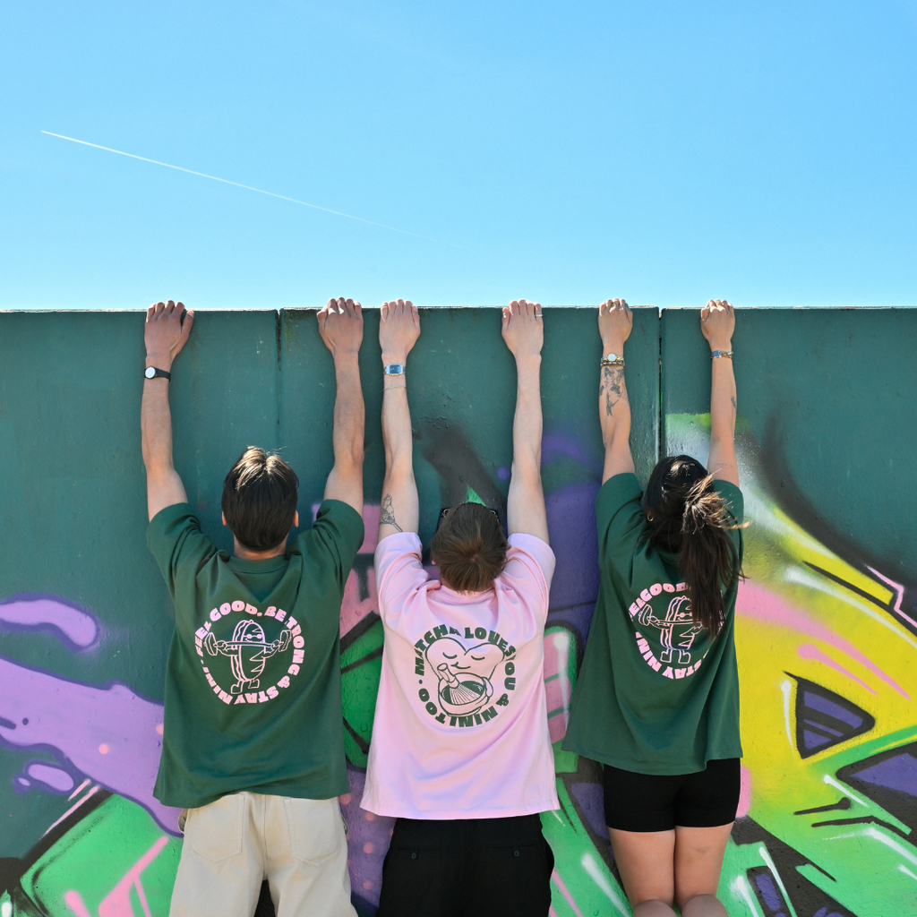 Drei Personen stehen vor einer Graffiti-Wand mit Armen nach oben, zwei grüne Café NINI Oversized T-Shirts mit pinkem „Feel Good Be Strong Stay NINI" Backprint und ein pinkes Shirt mit „Matcha Loves You & NINI Too" Backprint