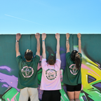 Drei Personen stehen vor einer Graffiti-Wand mit Armen nach oben, zwei grüne Café NINI Oversized T-Shirts mit pinkem „Feel Good Be Strong Stay NINI" Backprint und ein pinkes Shirt mit „Matcha Loves You & NINI Too" Backprint