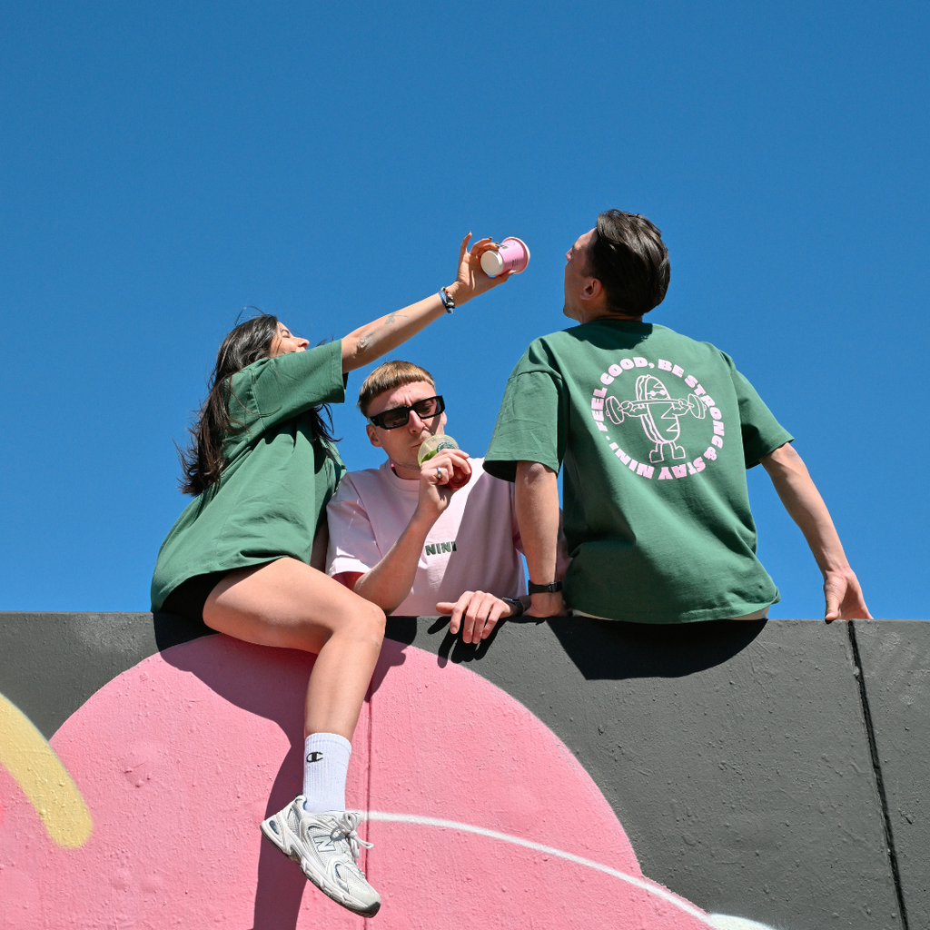Drei Personen sitzen auf einer Graffiti-Mauer vor blauem Himmel und tragen Café NINI Oversized T-Shirts in Grün und Pink, einer hält eine pinke NINI Keramiktasse, sommerliches Lifestyle Lookbook