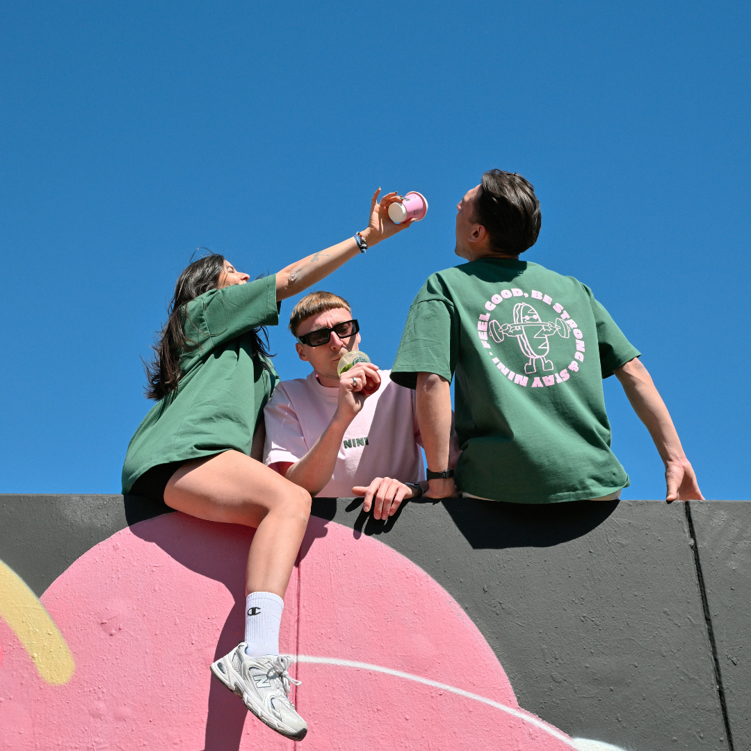 Drei Personen sitzen auf einer Graffiti-Mauer vor blauem Himmel und tragen Café NINI Oversized T-Shirts in Grün und Pink, einer hält eine pinke NINI Keramiktasse, sommerliches Lifestyle Lookbook