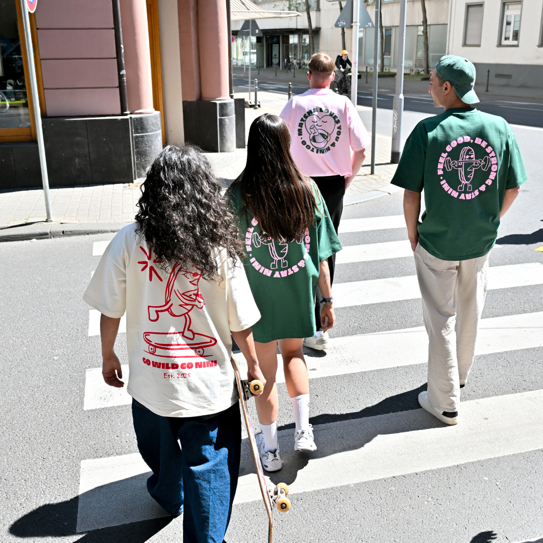 Vier Personen laufen über einen Zebrastreifen und tragen Café NINI Oversized T-Shirts, creme mit „Go Wild Go NINI Est. 2025" Backprint, grün und pink mit „Feel Good Be Strong Stay NINI" Backprint, urbaner Streetstyle mit Skateboard