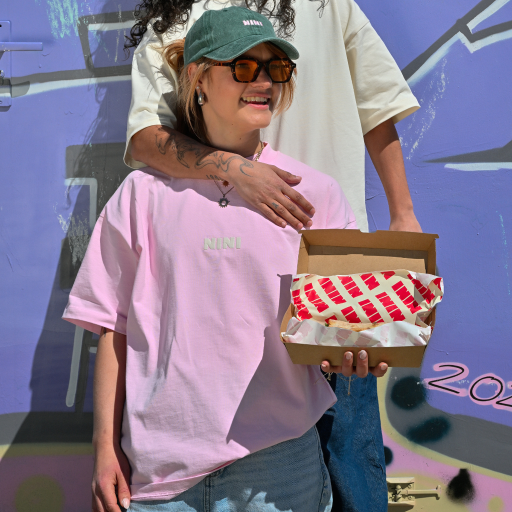Frau trägt Café NINI Oversized T-Shirt in Pink mit NINI Logo und hält eine Café NINI Verpackungsbox, im Hintergrund Person mit grüner NINI Cap und cremefarbenem Shirt, urbaner Streetstyle