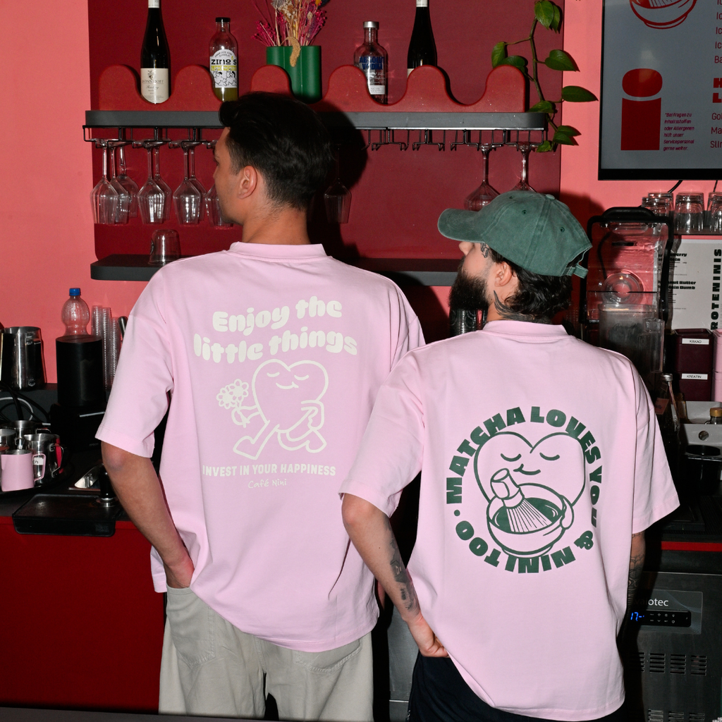 Zwei Baristas an der Kaffeebar tragen Café NINI Oversized T-Shirts in Pink, links Backprint „Enjoy the Little Things – Invest in Your Happiness", rechts Backprint „Matcha Loves You & NINI Too" mit grüner NINI Cap