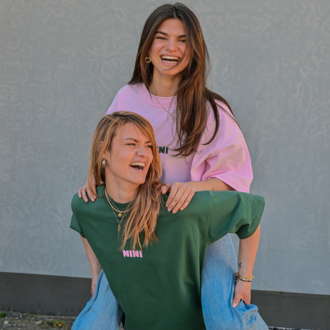 Zwei lachende Frauen tragen Café NINI Oversized T-Shirts in Pink und Grün mit pinkem NINI Logo, fröhliches Lifestyle Lookbook Duo Shot im Freien
