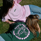 Zwei Frauen liegen auf einer Wiese und tragen Café NINI Oversized T-Shirts, pink mit grünem NINI Frontprint und grün mit pinkem „Feel Good Be Strong Stay NINI" Backprint, entspanntes Outdoor Lifestyle Lookbook

