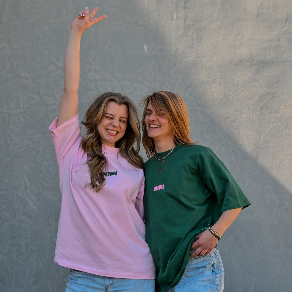 Zwei lachende Frauen tragen Café NINI Oversized T-Shirts in Pink und Grün mit pinkem NINI Frontprint, fröhliches Outdoor Lifestyle Lookbook
