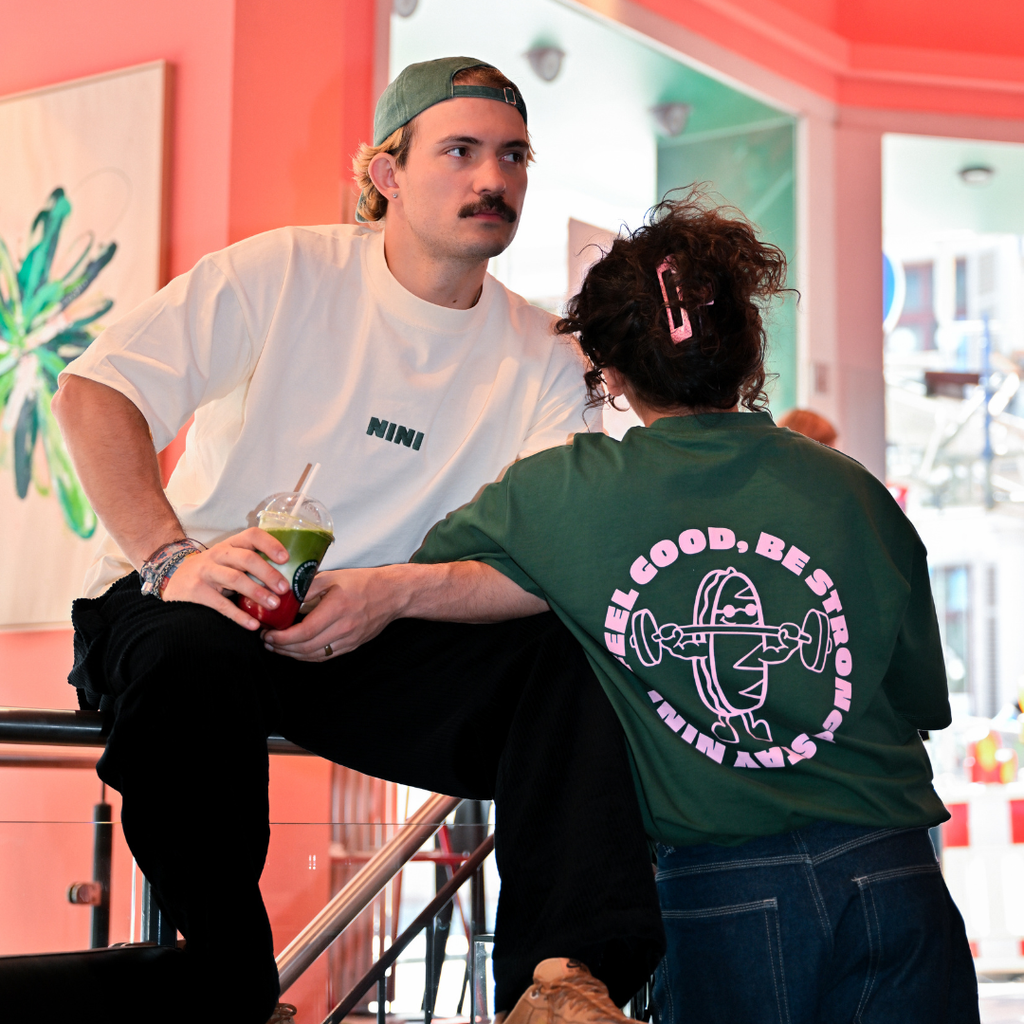 Mann und Frau im Café NINI tragen Oversized T-Shirts, weißes Shirt mit grünem NINI Frontprint und grünes Shirt mit pinkem „Feel Good Be Strong Stay NINI" Backprint, Mann hält grünen Matcha Drink, entspanntes Instore Lifestyle Lookbook