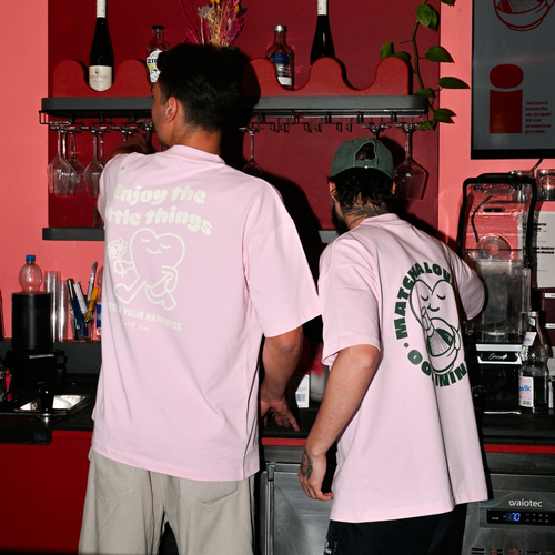 Zwei Baristas tragen Café NINI Oversized T-Shirts in Pink an der Kaffeebar - links „Enjoy the Little Things" Backprint, rechts „Matchalover NINI" Runddruck auf dem Rücken
