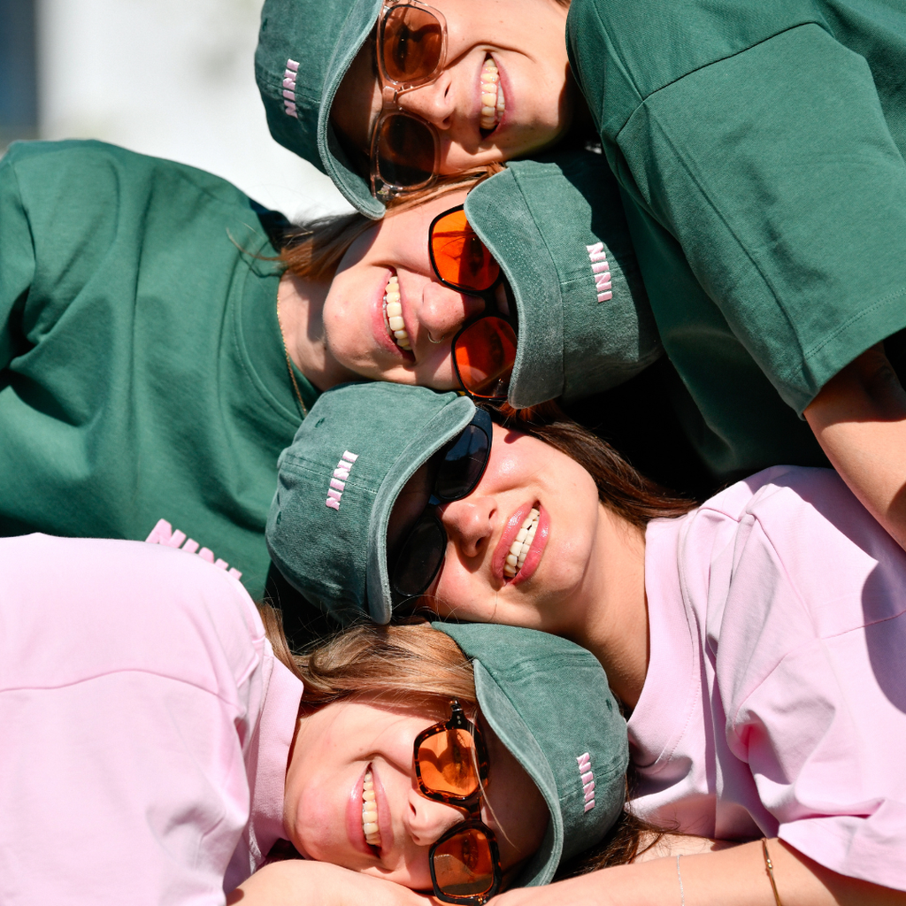 Vier lachende Personen tragen grüne Café NINI Dad Caps im Vintage Wash Look mit pinker NINI Stickerei und grüne NINI T-Shirts, fröhliches Gruppen Lifestyle Lookbook im Freien