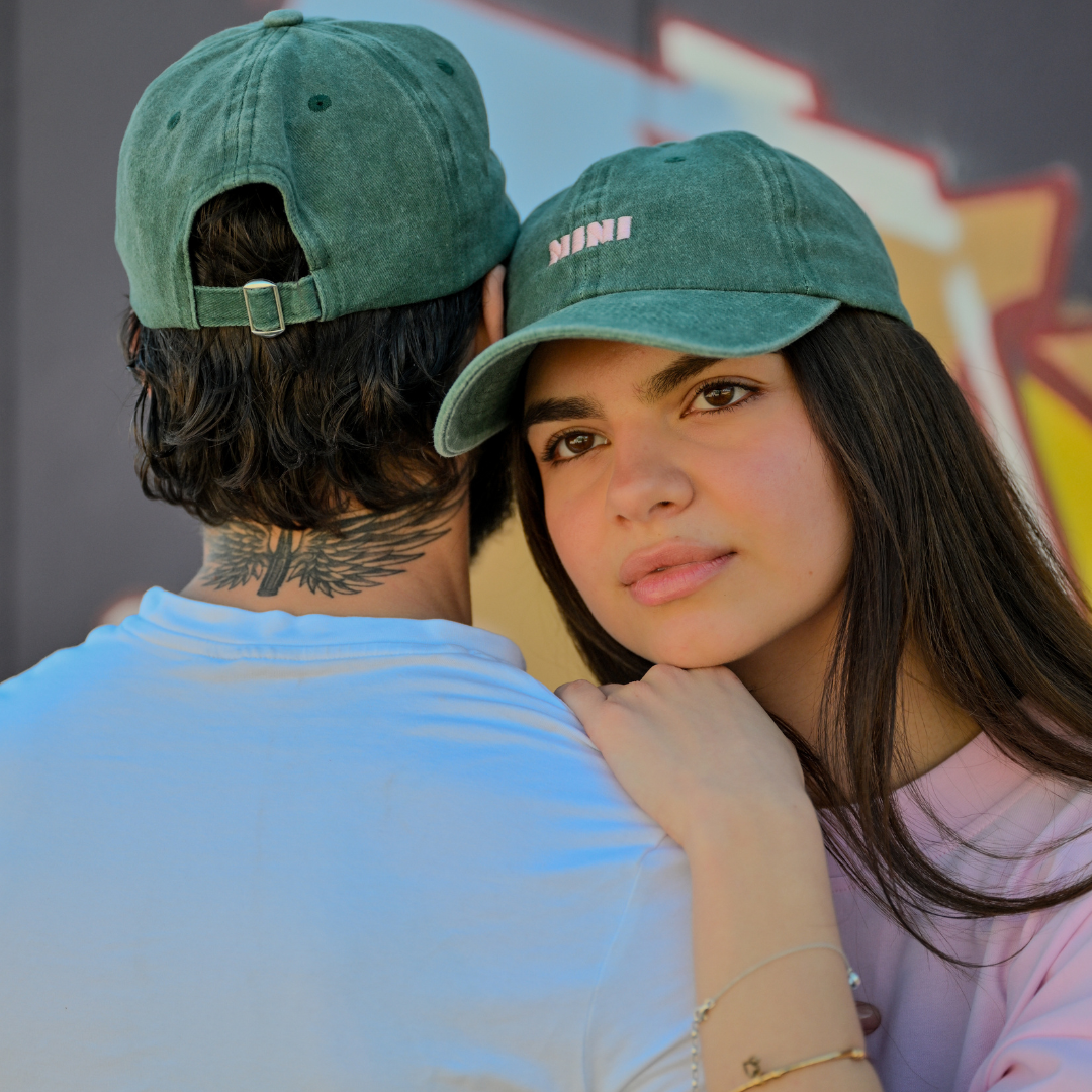 Zwei Personen tragen grüne Café NINI Dad Caps im Vintage Wash Look mit pinker NINI Stickerei, Front und Rückansicht, urbanes Duo Lifestyle Lookbook vor Graffiti-Wand