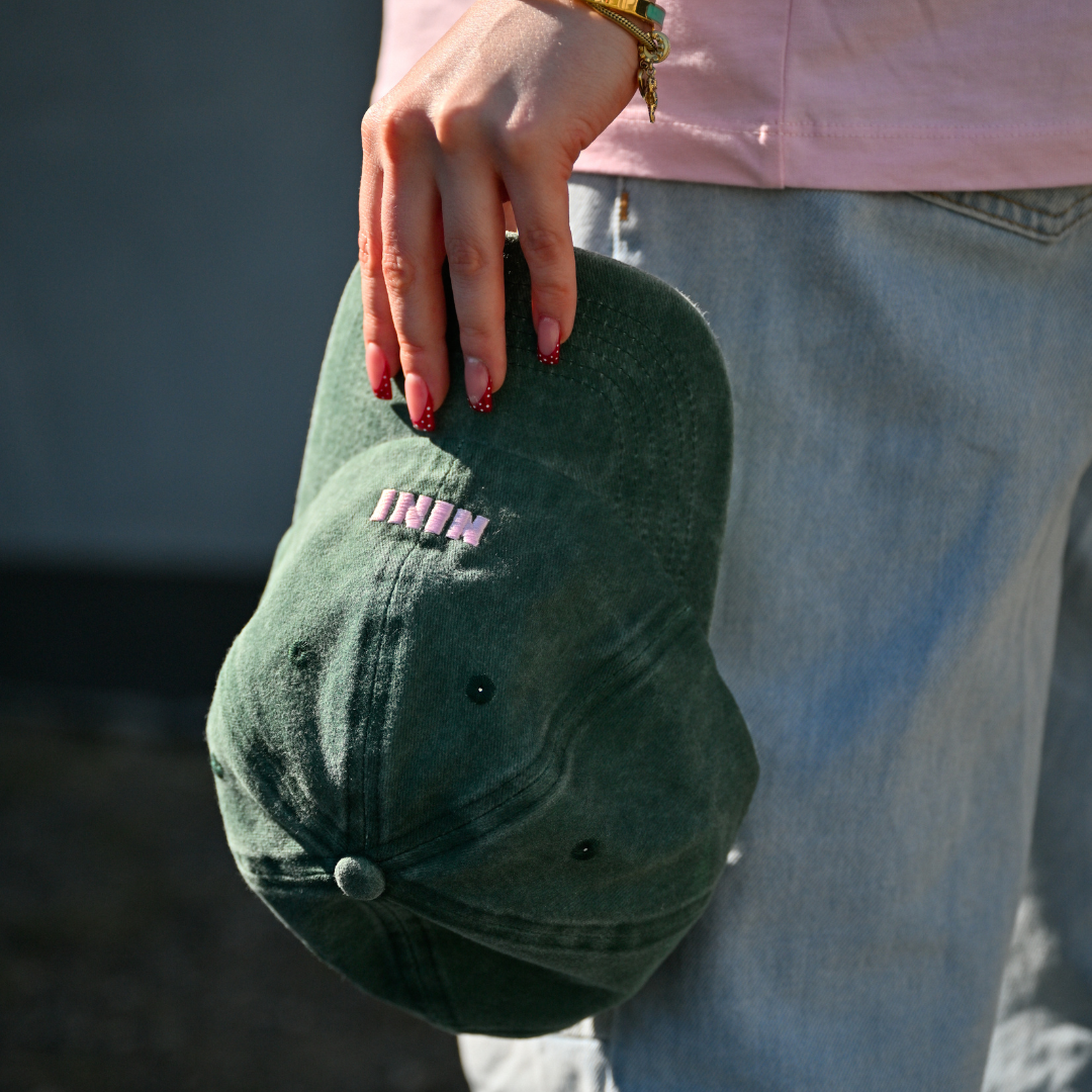 Grüne Café NINI Dad Cap im Vintage Wash Look mit pinker NINI Stickerei wird in einer Hand gehalten, Frau trägt pinkes NINI T-Shirt im Hintergrund, urbanes Outdoor Lifestyle Produktbild