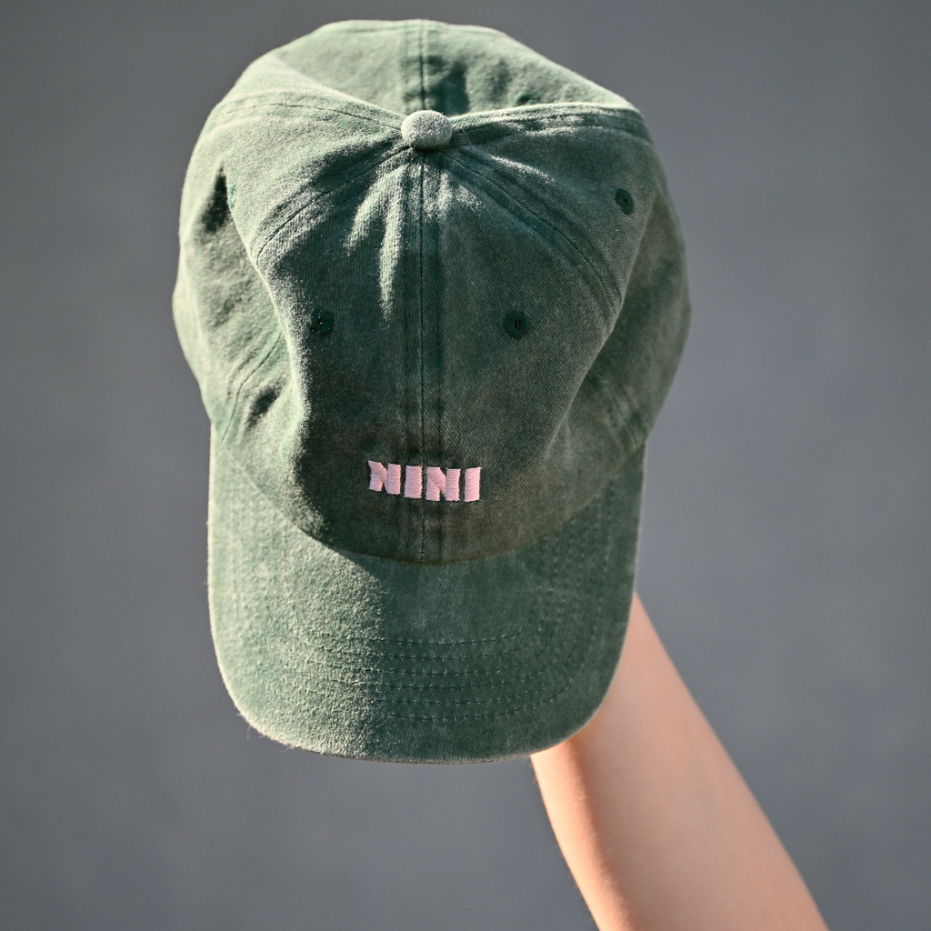 Grüne Café NINI Dad Cap im Vintage Wash Look mit pinker NINI Stickerei wird auf einer Hand präsentiert, Draufsicht Produktbild vor grauem Hintergrund