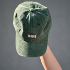 Grüne Café NINI Dad Cap im Vintage Wash Look mit pinker NINI Stickerei wird auf einer Hand präsentiert, Draufsicht Produktbild vor grauem Hintergrund