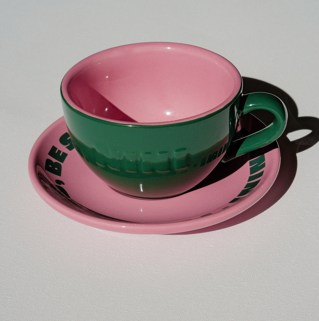 Café NINI Keramik Cappuccinotasse in Grün mit pinkfarbener Innenseite und pinker Untertasse mit eingeprägtem NINI Logo, seitliche Produktansicht auf weißem Hintergrund