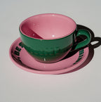 Café NINI Keramik Cappuccinotasse in Grün mit pinkfarbener Innenseite und pinker Untertasse mit eingeprägtem NINI Logo, seitliche Produktansicht auf weißem Hintergrund
