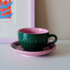 Grüne Café NINI Keramik Cappuccinotasse mit pinkfarbener Innenseite und pinker Untertasse steht auf einem Tisch vor einem bunten Kunstdruck, gemütliches Indoor Lifestyle Produktbild