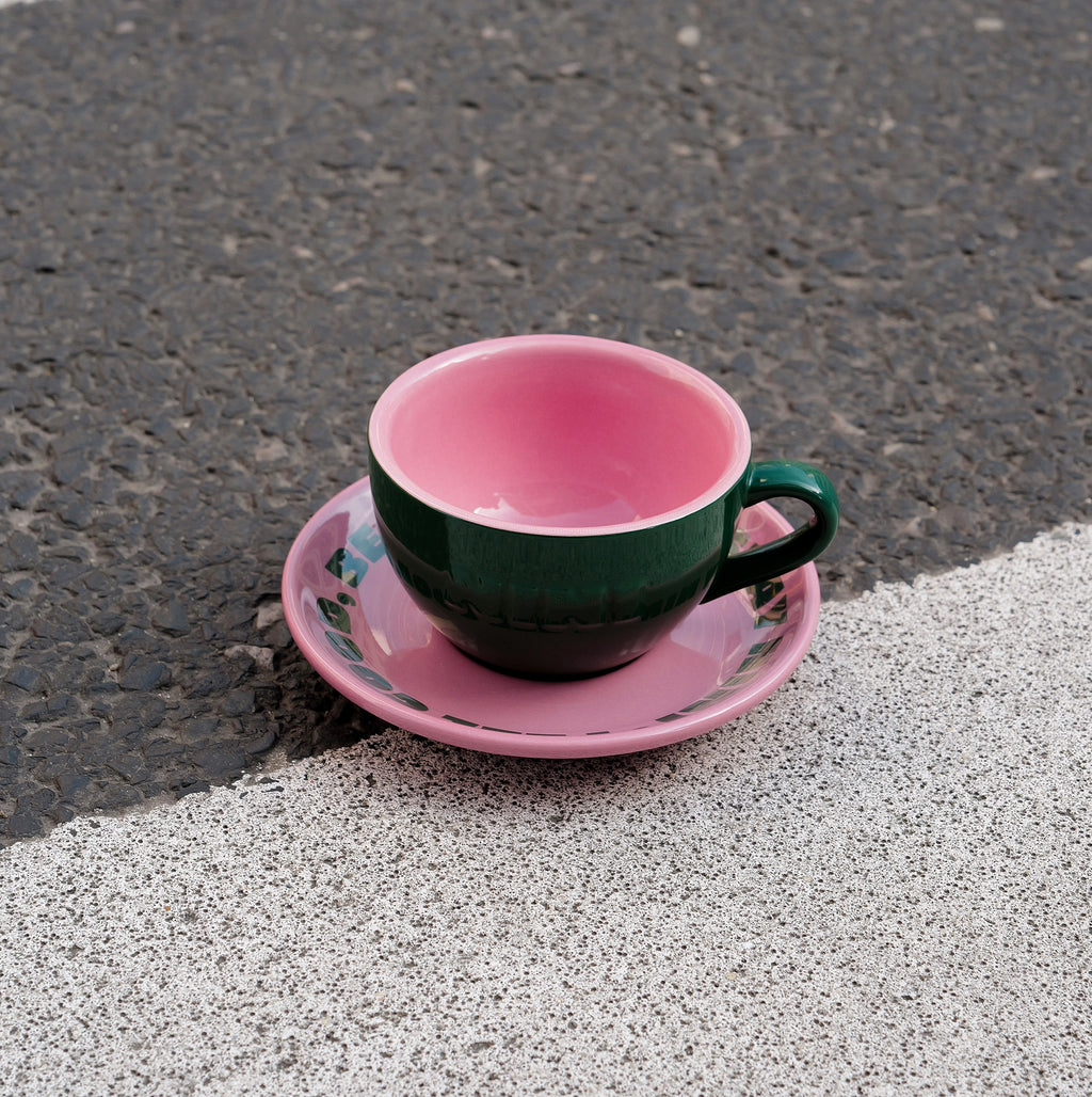 Grüne Café NINI Keramik Cappuccinotasse mit pinkfarbener Innenseite und pinker Untertasse steht auf Asphalt im Sonnenlicht, urbanes Outdoor Produktbild