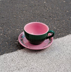 Grüne Café NINI Keramik Cappuccinotasse mit pinkfarbener Innenseite und pinker Untertasse steht auf Asphalt im Sonnenlicht, urbanes Outdoor Produktbild
