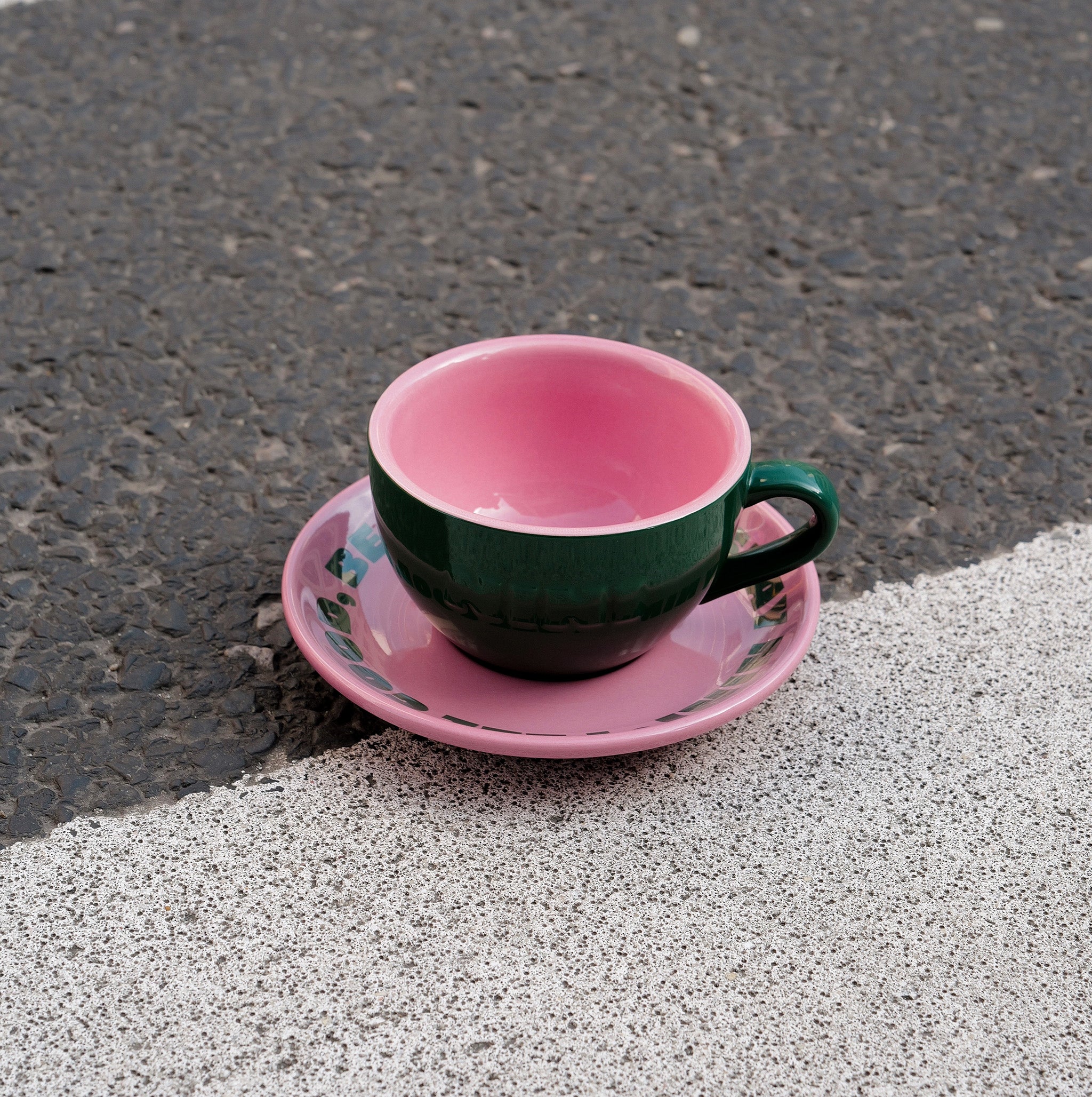 Grüne Café NINI Keramik Cappuccinotasse mit pinkfarbener Innenseite und pinker Untertasse steht auf Asphalt im Sonnenlicht, urbanes Outdoor Produktbild