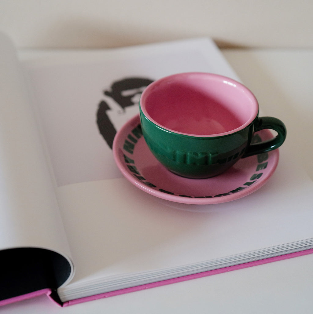 Grüne Café NINI Keramik Cappuccinotasse mit pinkfarbener Innenseite und pinker Untertasse liegt auf einem aufgeschlagenen Buch mit NINI Logo, gemütliches Lifestyle Produktbild