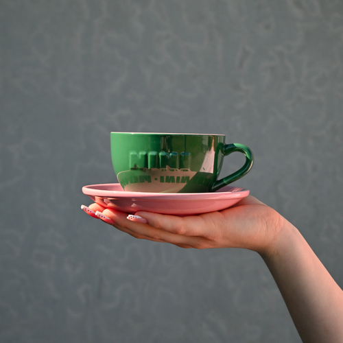 Grüne Café NINI Keramik Cappuccinotasse mit pinker Untertasse und eingeprägtem NINI Logo wird auf einer Hand präsentiert, Einzelproduktbild vor grauem Hintergrund