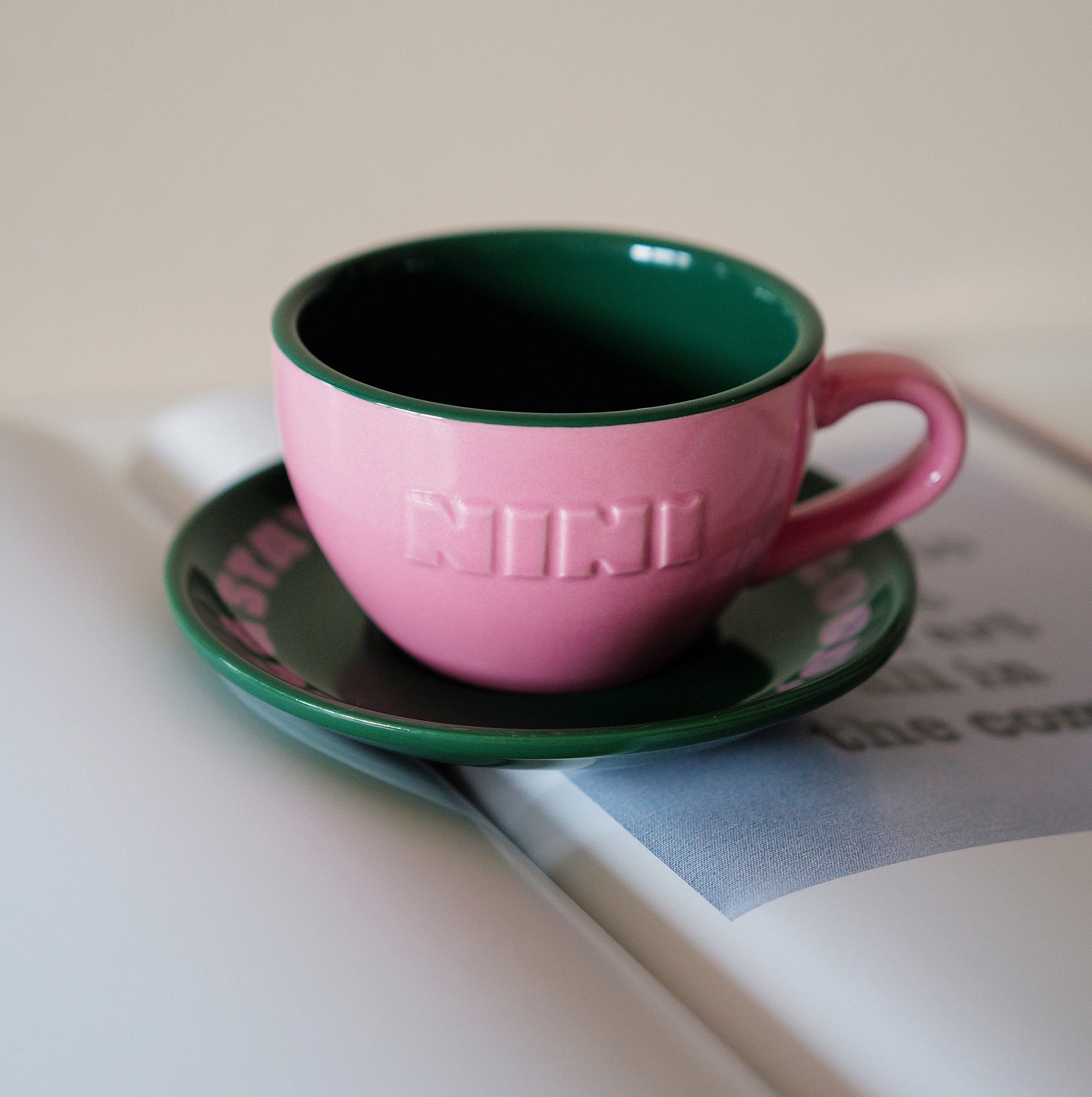 Pinke Café NINI Keramik Cappuccinotasse mit grüner Innenseite gefüllt mit Kaffee und grüner Untertasse liegt auf einem aufgeschlagenen Buch, gemütliches Indoor Lifestyle Produktbild