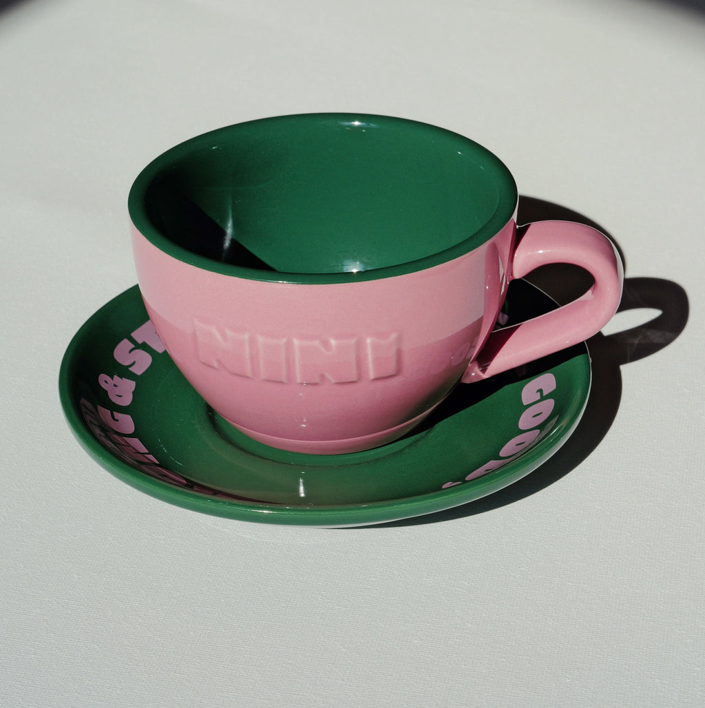 Café NINI Keramik Cappuccinotasse in Pink mit eingeprägtem NINI Logo und grüner Untertasse mit pinkem Schriftzug, seitliche Produktansicht auf weißem Hintergrund