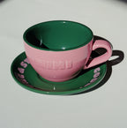 Café NINI Keramik Cappuccinotasse in Pink mit eingeprägtem NINI Logo und grüner Untertasse mit pinkem Schriftzug, seitliche Produktansicht auf weißem Hintergrund