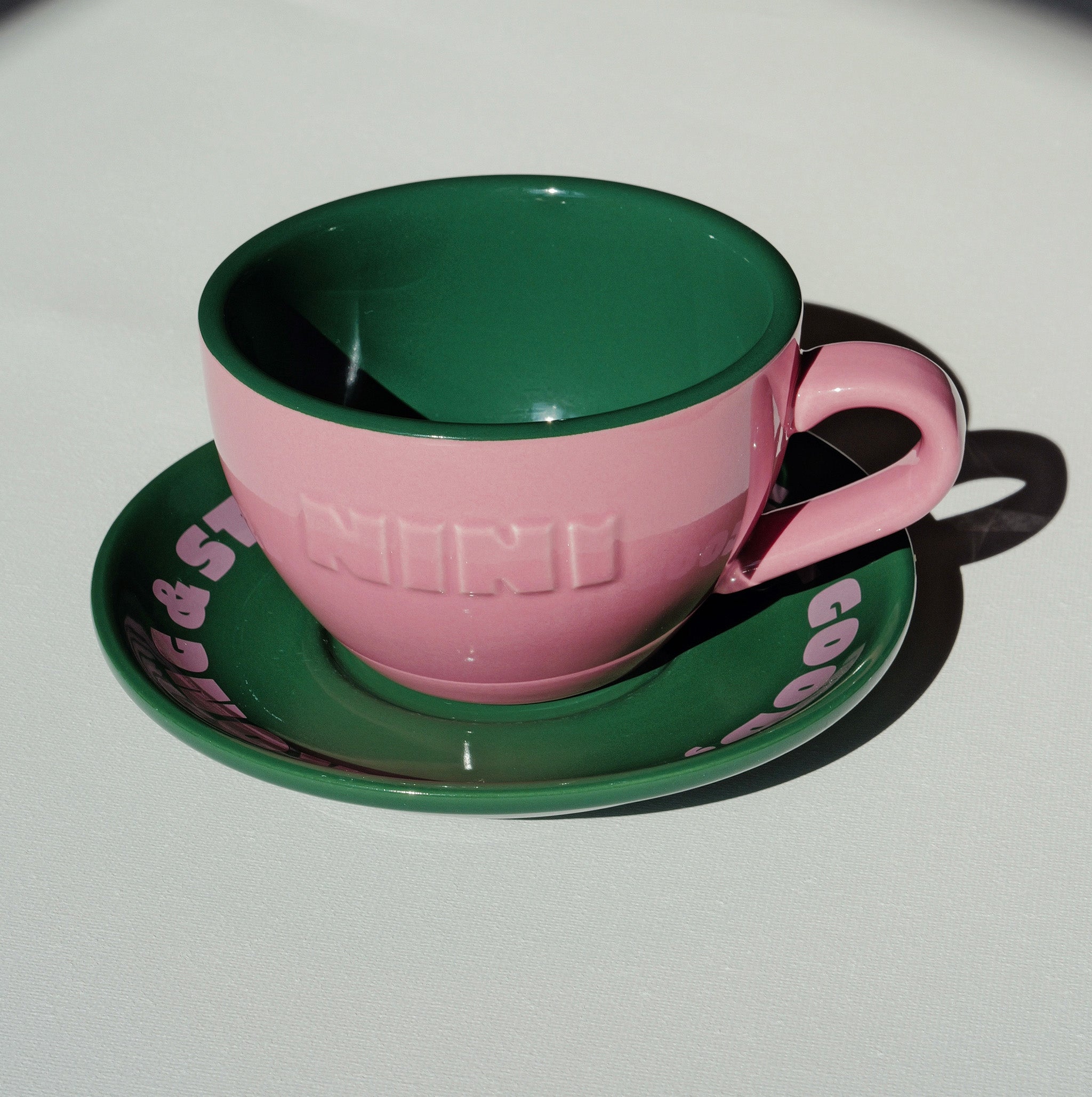 Café NINI Keramik Cappuccinotasse in Pink mit eingeprägtem NINI Logo und grüner Untertasse mit pinkem Schriftzug, seitliche Produktansicht auf weißem Hintergrund