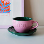 Pinke Café NINI Keramik Cappuccinotasse mit grüner Innenseite, eingeprägtem NINI Logo und grüner Untertasse steht auf einem Tisch vor einem bunten Kunstdruck, gemütliches Indoor Lifestyle Produktbild