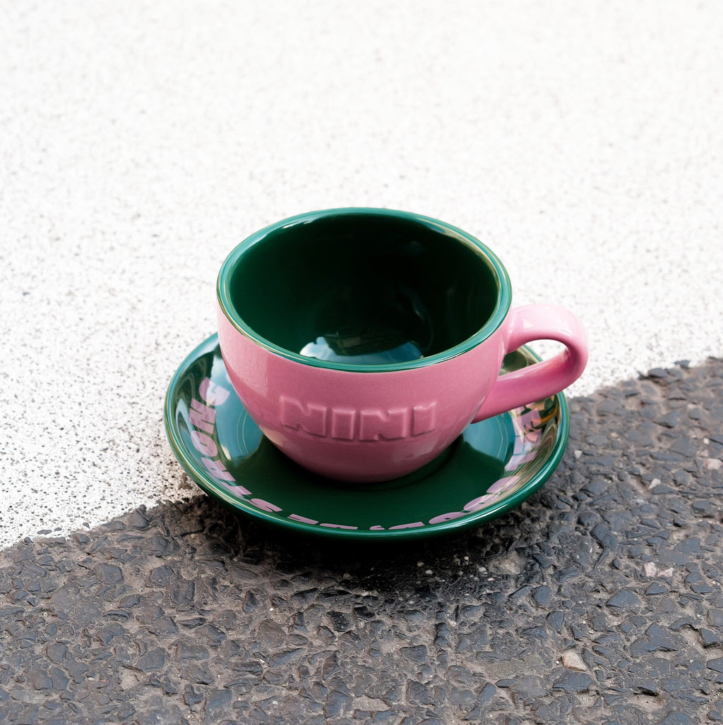 Pinke Café NINI Keramik Cappuccinotasse mit grüner Innenseite und grüner Untertasse steht auf Asphalt im Sonnenlicht, urbanes Outdoor Produktbild