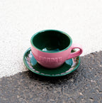 Pinke Café NINI Keramik Cappuccinotasse mit grüner Innenseite und grüner Untertasse steht auf Asphalt im Sonnenlicht, urbanes Outdoor Produktbild
