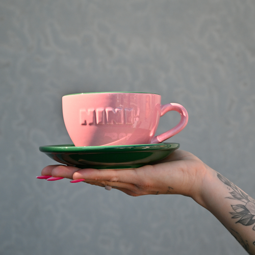 Pinke Café NINI Keramik Cappuccinotasse mit grüner Untertasse und eingeprägtem NINI Logo wird auf einer Hand präsentiert, Einzelproduktbild vor grauem Hintergrund