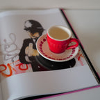 Rote Café NINI Keramik Espressotasse mit Espresso auf cremefarbener Untertasse liegt auf einem aufgeschlagenen Kunstbuch mit Street Art Illustration, kreatives Indoor Lifestyle Produktbild