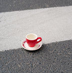 Rote Café NINI Keramik Espressotasse mit cremefarbener Innenseite und eingeprägtem NINI Logo auf cremefarbener Untertasse steht auf einem Zebrastreifen, urbanes Outdoor Produktbild