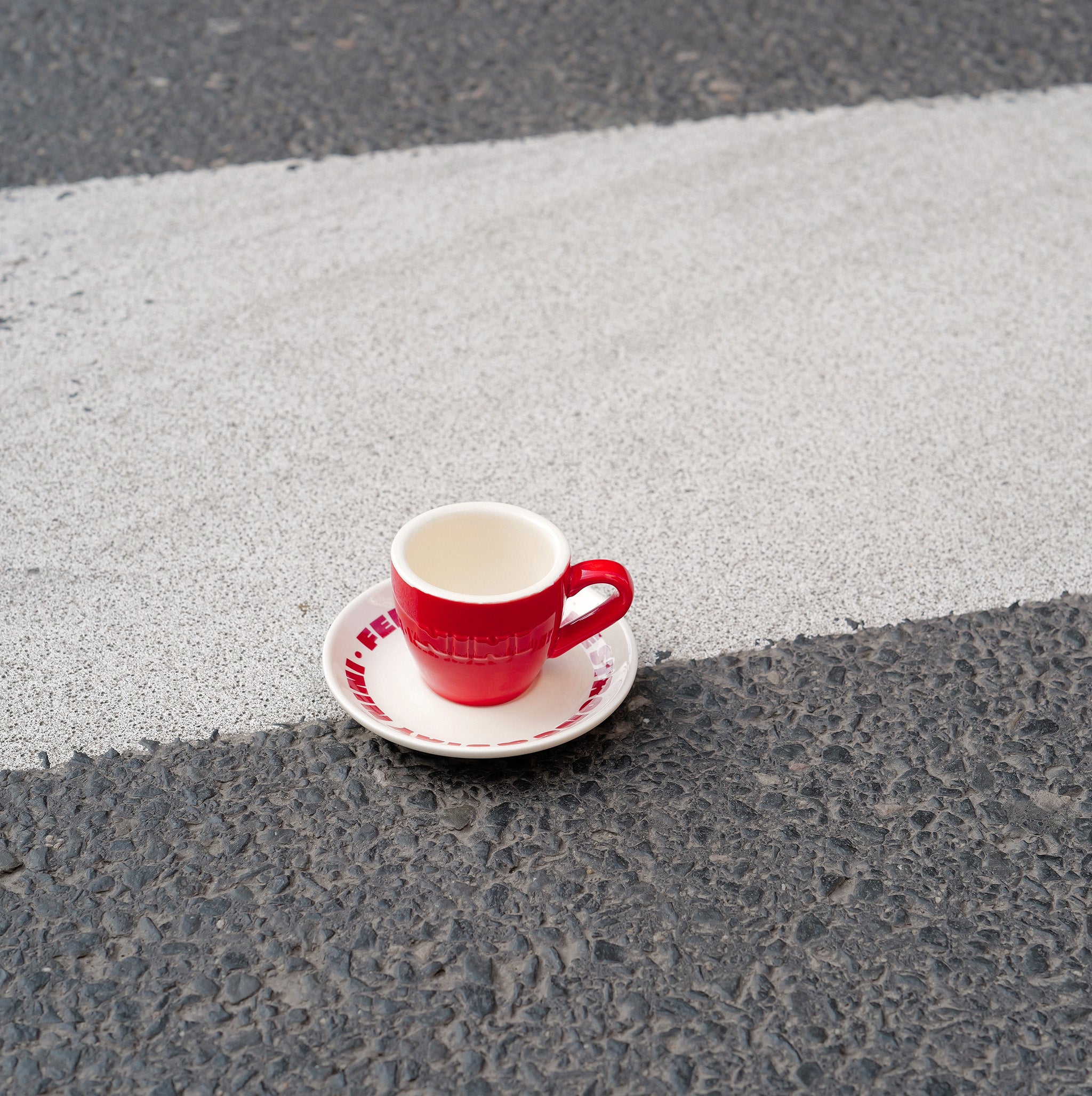 Rote Café NINI Keramik Espressotasse mit cremefarbener Innenseite und eingeprägtem NINI Logo auf cremefarbener Untertasse steht auf einem Zebrastreifen, urbanes Outdoor Produktbild