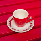 Rote Café NINI Keramik Espressotasse mit cremefarbener Innenseite und eingeprägtem NINI Logo auf cremefarbener Untertasse mit rotem „Feel Good Be Strong" Schriftzug, Produktbild auf rotem Lamellenuntergrund
